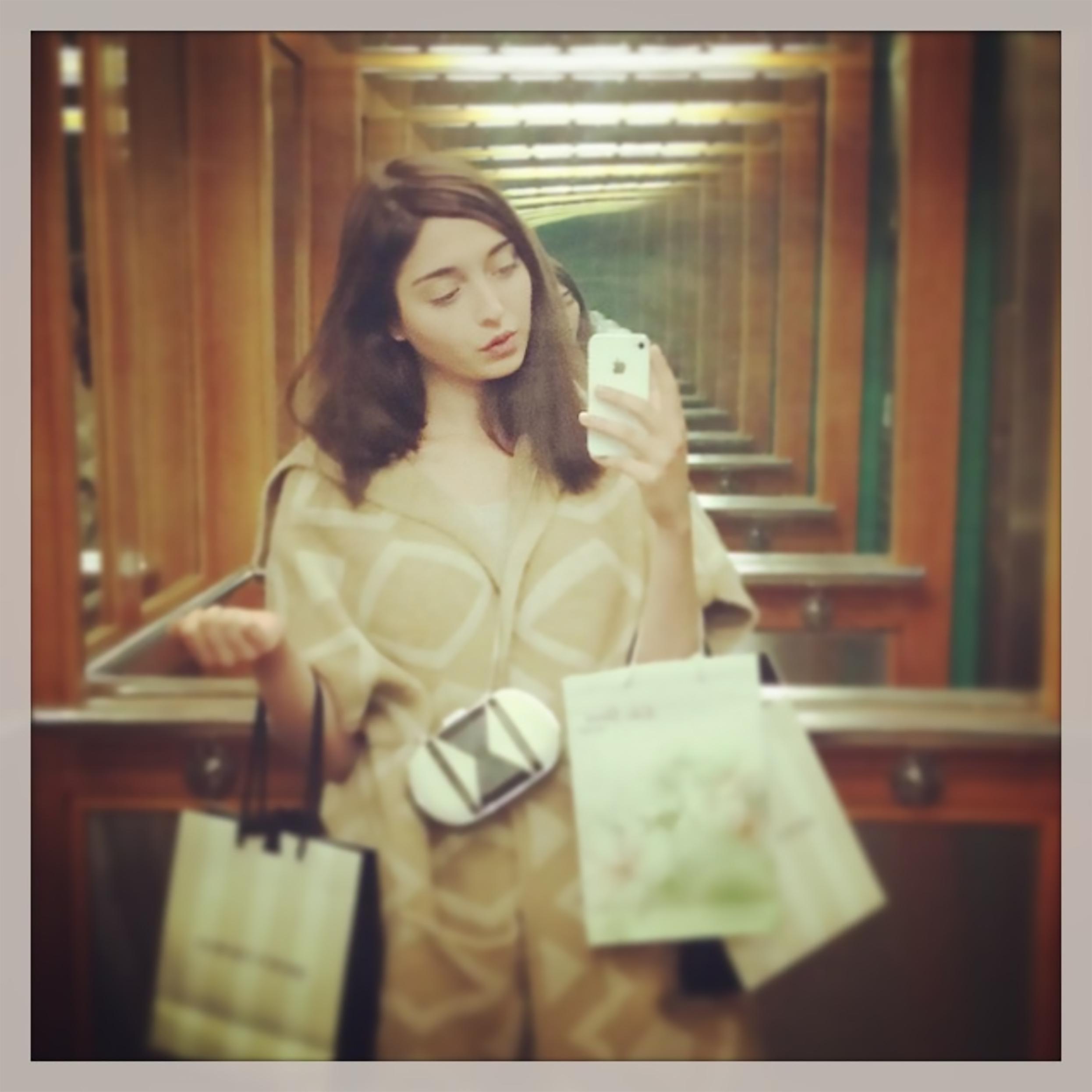 A brunette light-skinned woman takes a mirror selfie on her iPhone carrying shopping bags in the crooks of her arms.